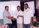 Neil Reece receives his ticket and monetary donation from Faizal Ally, Managing Director of A. Ally and Sons Limited in the presence of his coach Randolph Roberts (right).