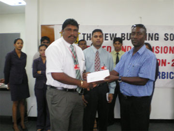  Operations manager of New Building Society Limited Anil Beharry hands over the sponsorship cheque to vice president of the Berbice Cricket Board of Control Mark Lyght as executives and staff of both entities look on.     