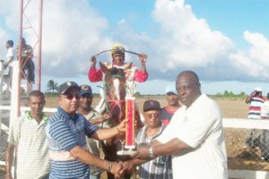 Public Relations Officer (PRO) of the Norman Singh Memorial Turf Club, Ian John right  presents the C&V Shipping Limited trophy to Chandrejeet Chattergoon who is the trainer of Kavinci (in background with jockey Yhap Drepaul astride).    