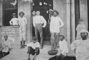 The Chinese proprietors of a village shop stand in the doorway surrounded by their customers, 1909