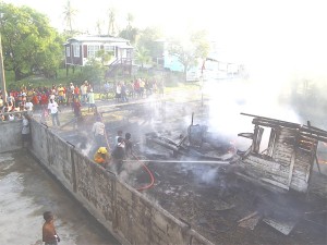 A few residents assist fire-fighters to put out flames while others look on. A fire, suspected to have been electrical in origin, destroyed this Industry, East Coast Demerara home of a family of four yesterday afternoon. (See story on page 11.) (Gaulbert Sutherland photo) 