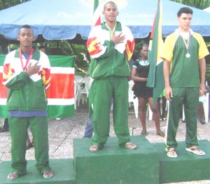 Suriname’s Marcelino Richaards (center) proudly sings his country’s national anthem after taking gold in the Boys 100 metre butterfly ahead of Guyana’s Nial Roberts (right) and Heneef Amatngalim (left) of Suriname. (Rawle Toney Photo)  