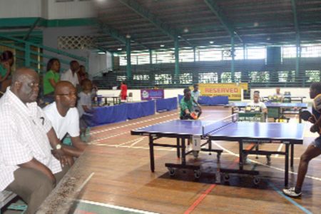 In this composite photo, Commissioner of Police Henry Greene (left), pays rapt attention to the finals of the Under-12 category of the National Sports Commission table tennis tournament between  Nigel Bryan, (seen returning the ball) and Joel Alleyne at the Cliff Anderson Sports Hall. (Orlando Charles photos)