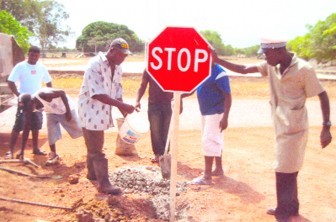 Region 9 Road Safety Council Chairman Terrence Boston (with bucket) and a constable of the police traffic department in Lethem erect a stop sign with the help of volunteers. 