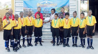 Redeemer Primary School’s football team display their medals as Petra Organisation Co-director Troy Mendonca collects a sponsorship cheque from a representative of Star Party Rental while Coach Johnny Barnwell (far left) and another representative (first, right back row) look on. 