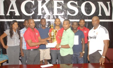 Retrieve Unknowns captain Travis Adolph (right in the foreground) collects the winning trophy and his team’s $250,000 first prize from ANSA McAl representative Jamal Douglas while other supporters look on.  
