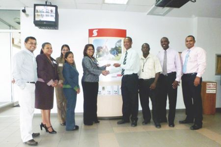 Scotiabank Personal Banking Manager, Ann Marie Singh (fourth from left) presents a cheque to AAG President Aubrey Hutson (fourth from right) as Coach, Mark Scott; Scotiabank Branch Manager, Matthew Langevine (second from right) and other bank employees look on.
