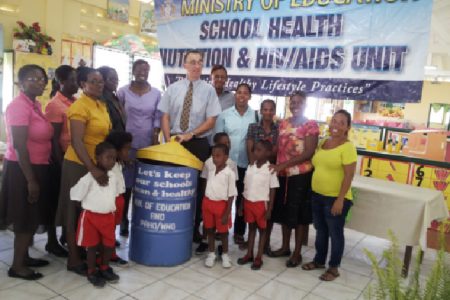 Adriannas Vlugman, Senior Advisor at PAHO (centre) along with Delma Nedd, Permanent Secretary of the Ministry of Education (to his left) pose with one of the bins and teachers, students and parents of the Enterprise Nursery School.