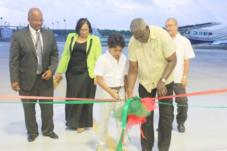 A young man assisting Minister of Works Robeson Benn in cutting the ribbon to commission the Wings Aviation Limited hangar at Ogle International Airport. Looking on (from left to right) are CEO of Wings Aviation Ronald Reece, his wife Roxanne who is the General Manager and Director General of the Guyana Civil Aviation Authority Zulficar Mohamed.