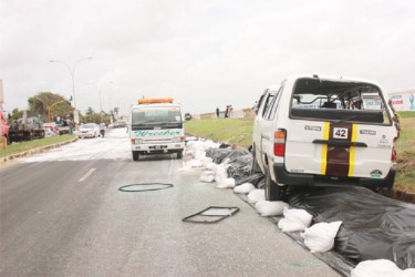 The minibus perched atop sandbags on the Rupert Craig Highway after it had toppled several times. In the background is the foamed section of the road where the oil had spilled. (Photo by Arian Browne) 