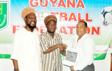 GFCA President Wayne Dover (centre) hands over the national programme to GFF VP of Technical and Tactical Development Ivan Persaud (right) while GFCA General Secretary Sampson (left) Gilbert looks on.