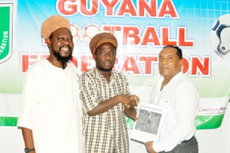 GFCA President Wayne Dover (centre) hands over the national programme to GFF VP of Technical and Tactical Development Ivan Persaud (right) while GFCA General Secretary Sampson (left) Gilbert looks on.
