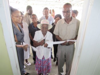 Dr Abbigale Jeffrey, the operations officer of the Mon Repos Health Centre and Joesph Hamilton, Parliamentary Secretary for the Ministry of Health, assist as an elderly member of the community cuts the ribbon for the reopening of the refurbished facility. 
