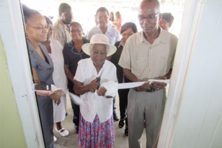 Dr Abbigale Jeffrey, the operations officer of the Mon Repos Health Centre and Joesph Hamilton, Parliamentary Secretary for the Ministry of Health, assist as an elderly member of the community cuts the ribbon for the reopening of the refurbished facility. 
