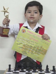 Little Sparsh Bisht from India pictured with a chess trophy and his chess pieces. Sparsh began playing chess at three years and ten months of age. He was born in 2009 and was a participant in two national chess tournaments in India.
