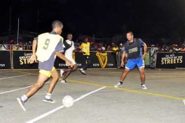 Roy Cassou (no.9) of Albouystown-B on the attack as Elroy Parks (black vest/far right) of Stevedore Housing Scheme try to defend during their semi-final matchup. (Orlando Charles photo) 
