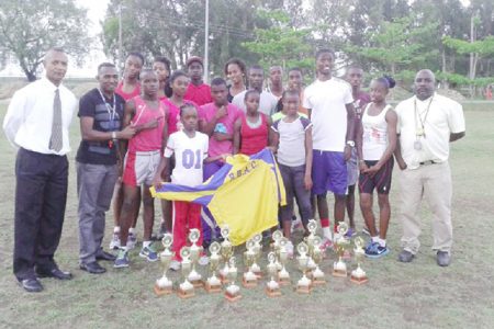 Athletes of the Running Brave Athletic Club pose with the hardware they earned at the recently concluded Hampton Games. They are flanked by coaches, Sham Johnny and Julian Edmonds along with president of the club and the AAG, Aubrey Hutson.
