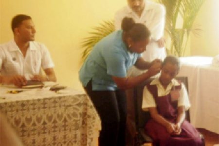 Minister of Health Dr Bheri Ramsaran (standing) looks on as a nurse fits a child with a hearing aid.