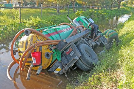 This Cevon’s Waste Management truck went overboard on Friday in a trench near the University of Guyana. According to the driver, he had been “pushed off” the road by two cars. In this Arian Browne photo, two employees of the company hasten to remove the vehicle’s battery and other parts to prevent further damage.
