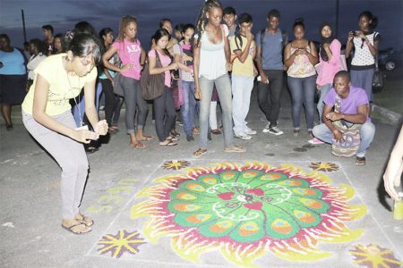 A participant adding light to the University of Guyana’s Economics Society’s Rangoli as other students view the creation. (Photo by Arian Browne)