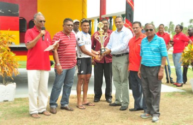 Last year’s overall winner Alanzo Greaves receives the champion trophy from Minister of Sport Dr. Frank Anthony in the presence of race organizer Hassan Mohammed, Director of Sport Neil  Kumar and other stakeholders. (Orlando Charles photo) 