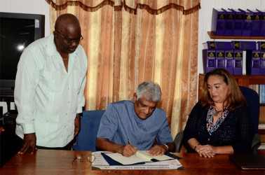 GECOM Chairman Dr Steve Surujbally signing the document in the presence of Lisa Shoman (right) and Chief Election Officer Keith Lowenfield. (GECOM photo) 