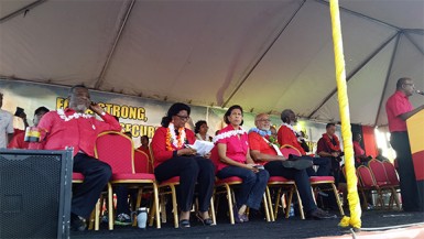 Prime Minister Samuel Hinds (left) relaxes on the stage holding a PPP/C flag-covered cup while Prime Ministerial Candidate Elisabeth Harper (second from left)  looks over her notes. Former President Bharrat Jagdeo is at the lectern.