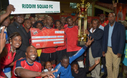 Riddim Squad’s captain Lylton Ramsay collects the championship trophy from GFA President Clifton Hicken while members and supporters of the team celebrate following their win over Western Tigers in the GFA/Banks Beer Cup final at the Providence National Stadium Friday