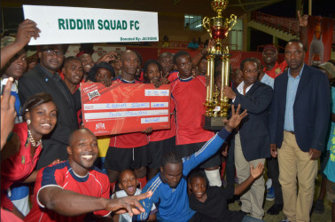 Riddim Squad’s captain Lylton Ramsay collects the championship trophy from GFA President Clifton Hicken while members and supporters of the team celebrate following their win over Western Tigers in the GFA/Banks Beer Cup final at the Providence National Stadium Friday