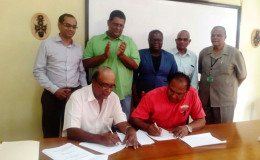 CEO of NAREI, Dr. Oudho Homenauth (sitting at left) signing for NAREI while Anand James signs for AMU Agro-Processors. NAREI Chairman  Major General (ret’d) Joseph Singh is standing at right.