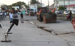 A rollerblader makes use of the recently cleared “skating rink” along the Merriman Mall strip between Cummings Street and Orange Walk, while contractor Cevon’s waste management removes waste from the site. Stabroek News was able to observe four skips of waste being removed from the area last evening. (Photo by Keno George) 