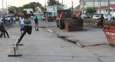 A rollerblader makes use of the recently cleared “skating rink” along the Merriman Mall strip between Cummings Street and Orange Walk, while contractor Cevon’s waste management removes waste from the site. Stabroek News was able to observe four skips of waste being removed from the area last evening. (Photo by Keno George) 