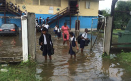 Students and a teacher with their footwear in their hands manoeuvre  through the flooded school yard. 