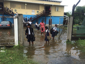 Students and a teacher with their footwear in their hands manoeuvre through the flooded school yard. 