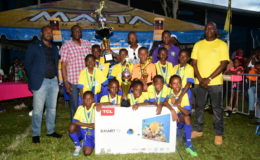 St. Angela’s captain Antwoine Vincent (3rd from left) displaying the championship trophy alongside his teammates following their hard-fought win over St. Agnes in the Courts Pee Wee Football final while GFF President Wayne Forde (left), Courts Financial Controller  Neil Boucher (2nd from left), Head-Coach Oscar Payne (centre), Courts Marketing Director Pernel Cummings (2nd from right) and Banks DIH Limited Malta Brand Manager Clayton McKenzie (right) look on 