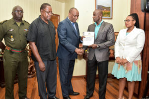 Minister of State,  Joseph Harmon (centre)  receives the Report from Brigadier (Retired) Bruce Lovell as Lieutenant Colonel Denzel Carmichael (left), former Assistant Commission of Police, . Winston Cosbert (second from left) and Ms. Christine Bailey look on.  (Ministry of the Presidency photo)