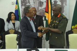 Brigadier Patrick West (right) hands over a ceremonial “coin” to University of Guyana Vice Chancellor, Ivelaw Griffith (DPI photo)
