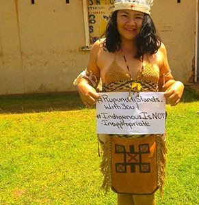 A young woman of Amerindian heritage bearing a placard which states: “#RupununiStandsWithYou” and “#IndigenousIsNotInappropriate”