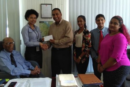 Maurice Solomon (sitting) looks on as Charmaine Walker (left standing) hands over the sponsorship cheque over to LGC President Aleem Hussain in the presence of some staff members of Maurice Solomon and Company.
