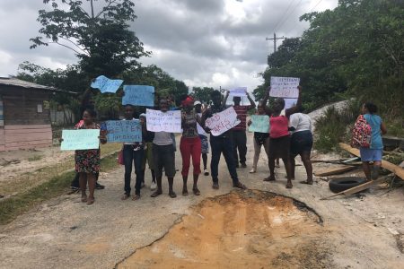 Parents and residents of Kuru Kuru, Soesdyke-Linden Highway, with their placards yesterday, standing in front of the first pothole at the start of the access road.
