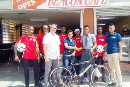 Proprietor of Beacon Café Ramdeo Kumar (front row extreme left), Accounts Clerk of Beacon Café Ateeyyea Ali presents Jodel Fernandes with his new bicycle in the presence of Feaz Mustapha of Beacon Café (front row extreme right) and members of the Beacon Football Club