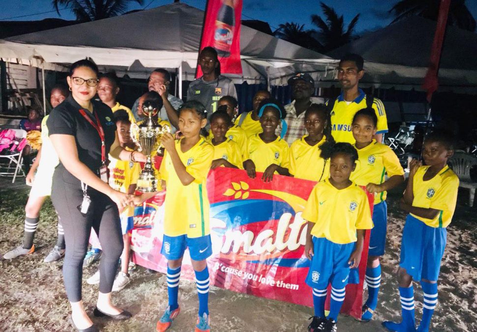 Champions At-Last-West Ruimveldt captain receiving the championship trophy from the Smalta Brand Representative in the presence of her team-mates, after defeating North Georgetown in the finale of the Girls Pee Wee Football Championship at the Ministry of Education ground, Carifesta Avenue 
