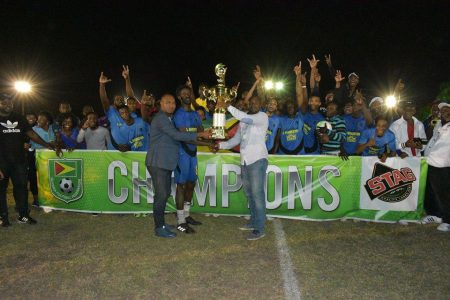 The victorious Den Amstel captain Kester Jacobs collecting the championship trophy from GFF President Wayne Forde [right] and Stag Brand Representative Nigel Worrell [left] in the presence of his celebrating team-mates
