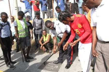 People look on as this policeman tries to lift this manhole cover near Tangerine Place in St Andrew in order to facilitate a search for a man who was shot in an alleged robbery attempt yesterday. 