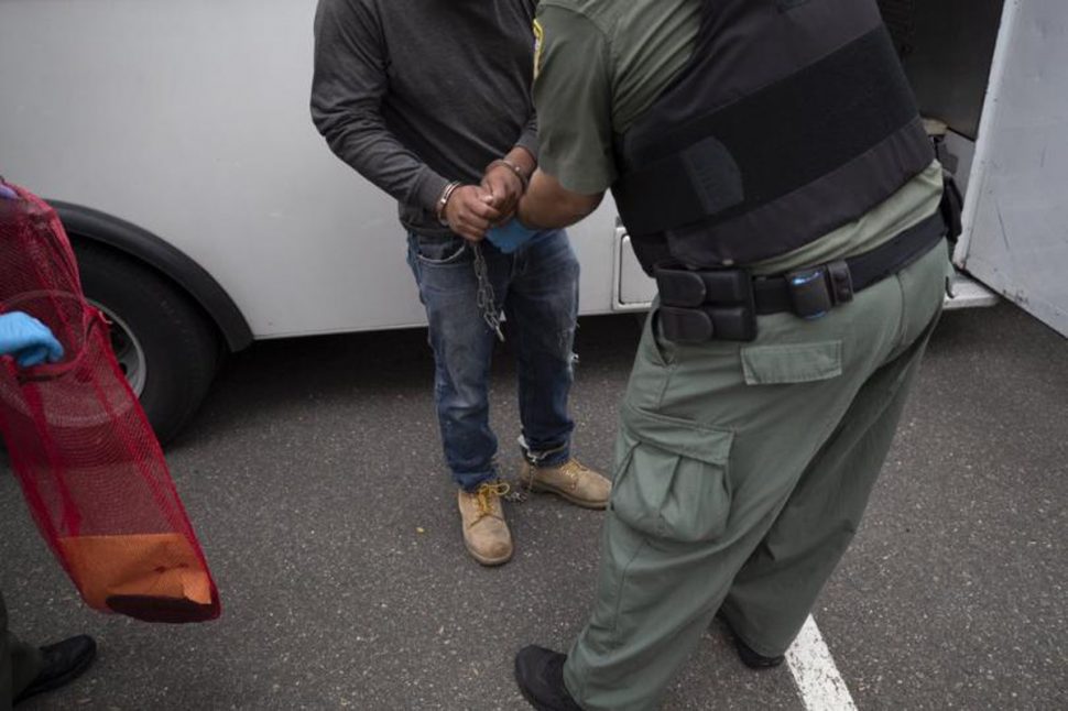 In this July 8, 2019, file photo, a US Immigration and Customs Enforcement officer transfers a man in handcuffs and ankle cuffs on to a van during an operation in Escondido, California. Advocacy groups and unions are pressuring Marriott, MGM, and others not to house migrants who have been arrested by US Immigration and Customs Enforcement agents, but the US government says it sometimes needs bed space, and if hotels don’t help, it might have to split up families.