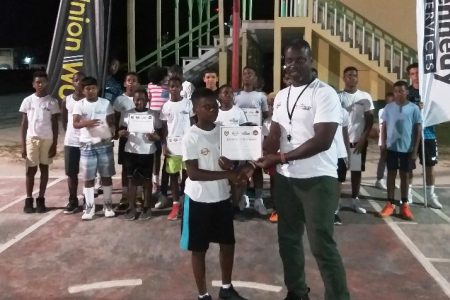 HBA head Junior Hercules presenting a certificate of participation to one of the players during the conclusion of the second  annual HBA Bartica Summer Camp in the presence of several other participants.