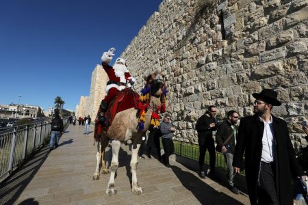 An Ultra-Orthodox Jewish man looks at Mr Issa Kassissieh as he rides a camel while wearing a Santa Claus costume during the annual Christmas tree distribution organised by the Jerusalem municipality in Jerusalem’s Old City on Dec 19, 2019.
