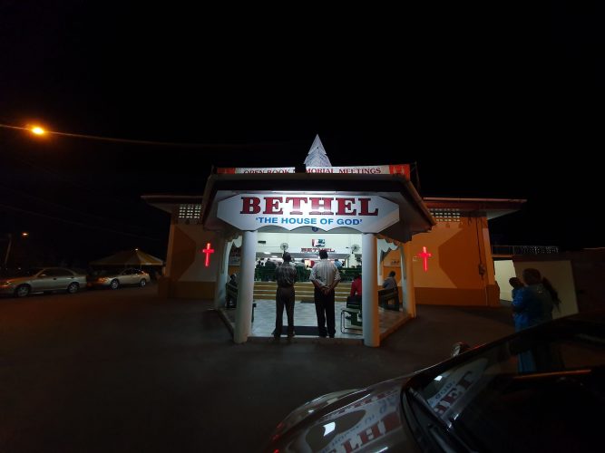 Two congregation members stand at the entrance to the Bethel ‘The House of God’ church during a service on Thursday night. The service was held in contravention of the current COVID-19 protocol which debars social gatherings of more than 10 people. Police showed up at the venue but took no action to stop the event. 