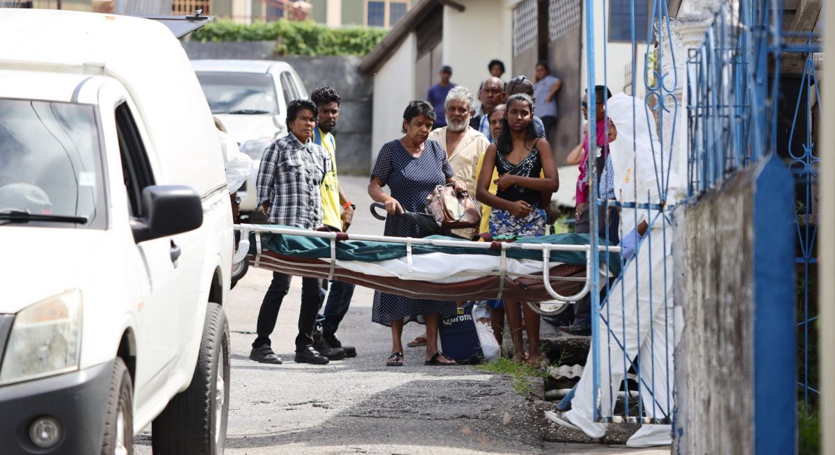 A sad Radhica Rampersad, left, and her son Krishna Seepersad look on as undertakers remove the body her daughter Susan Seepersad, after she was killed at her home in Rampersad Trace Debe, yesterday.