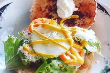 A toasted breakfast roll with sunny side eggs, lettuce, tomato, cucumber, mustard, ricotta cheese, and a dollop of sour cream. 